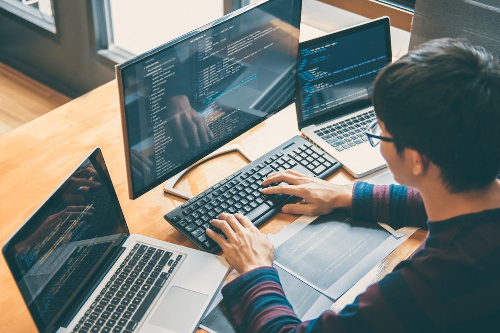 Homem sentado em uma mesa com três notebooks, programando