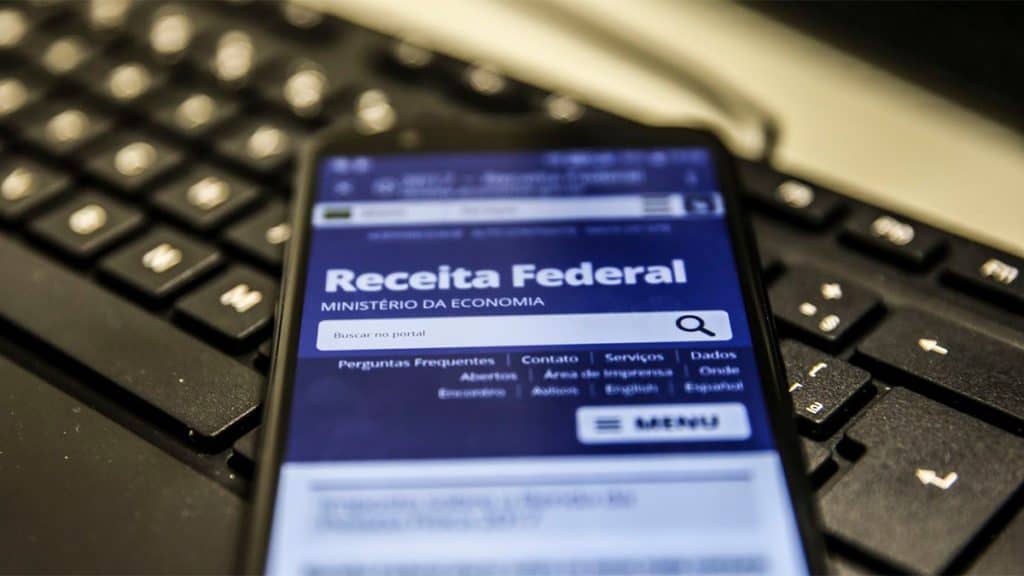 Celular com homepage da Receita Federal aberta em navegador; aparelho está sobre teclado de computador